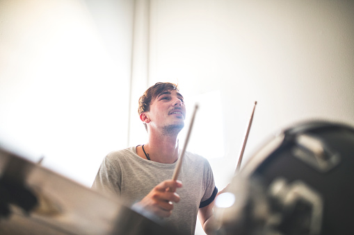 Young drummer practicing in recording studio. Rock musician is playing drum kit. He is sitting against wall.