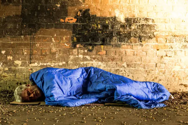 Low angle view of a homeless senior adult male, of caucasian ethnicity, lying down and sleeping rough outdoors in a subway tunnel. The floor of the dark tunnel is uncomfortable and covered with stone. Behind the homeless man is a brick wall. The man, who has white hair and white stubble on his face, is inside a blue sleeping bag, and we can only see his face. His eyes are closed, and he is sleeping. Lots of room for copy space.