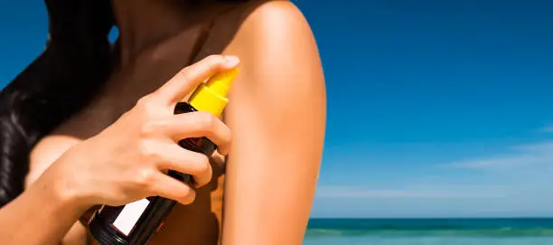 Close - up of Sexy asian woman are Spray Sunscreen , lotion or sunblock to body for prevent UV rays from sunlight Before the sun bathing in the summer or Vacation with sea and bluesky bckground