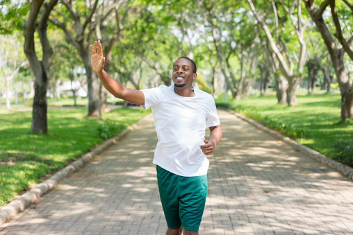 Friendly sporty black guy jogging in city park and greeting familiar sportsmen. Young Afro American man running outdoors and waving hand. Sport and communication concept