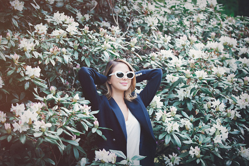 Young redhead girl in white sunglasses near flowers at springtime blossom garden.