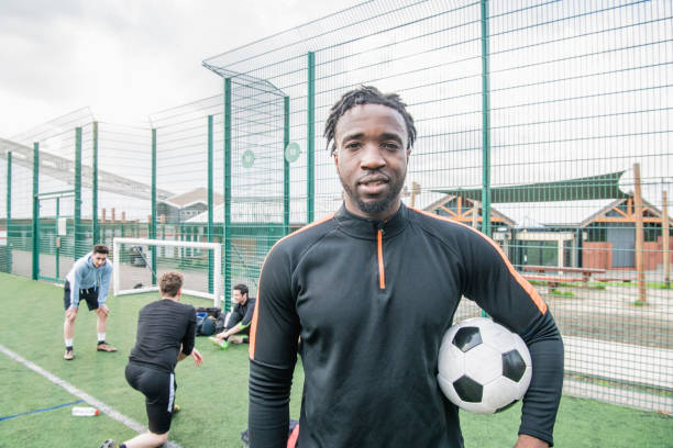 Portrait of young man holding football 19 year old man, african ethnicity ready to play football at urban all weather pitch, London soccer player photos stock pictures, royalty-free photos & images