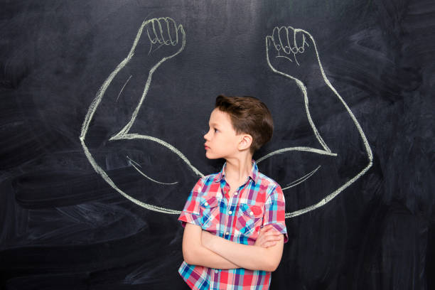 istock Portrait of serious kid looking on blackboard with drawn strong biceps 954144994