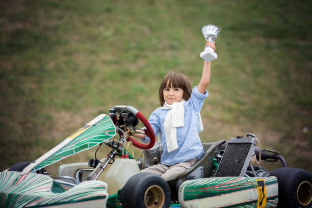 Happy little girl seated on a green bumper motorcycle, navigating a paved surface within an outdoor recreational setting. The motorcycle features a protective bumper circling its base, designed for safe and controlled movement. In the background, a row of red and white painted tires lines the edge of the pavement, acting as barriers.\nShot with Nikon D800