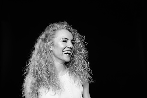 Close up studio portrait of a pretty curly blonde woman, wearing light dress, laughing and looking to the side, against plain studio background
