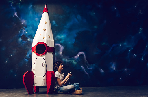 Boy is sitting near toy rocket on space background and listening to music with smart phone in hands.