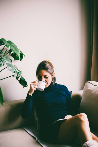 istock Beautiful young woman sitting on sofa, looking at tablet screen and drinks coffee (toned image, selective focus) 954264498