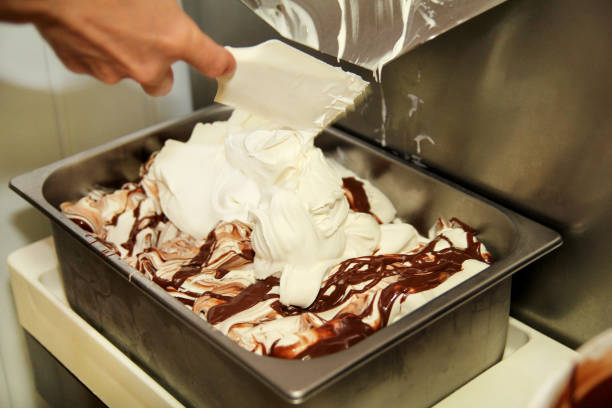 istock Female worker is working on ice cream maker machine. Producing vanilla ice cream with chocolate dressing and it falls into steel container. Industrial preparation of creamy ice cream. Dessert, sweets. 954276622