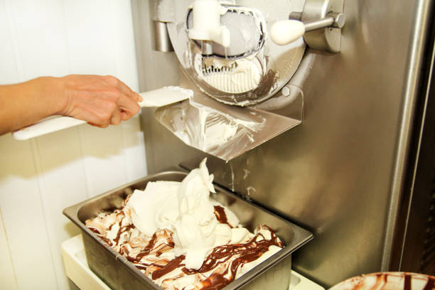 istock Female worker is working on ice cream maker machine. Producing vanilla ice cream with chocolate dressing and it falls into steel container. Industrial preparation of creamy ice cream. Dessert, sweets. 954276636
