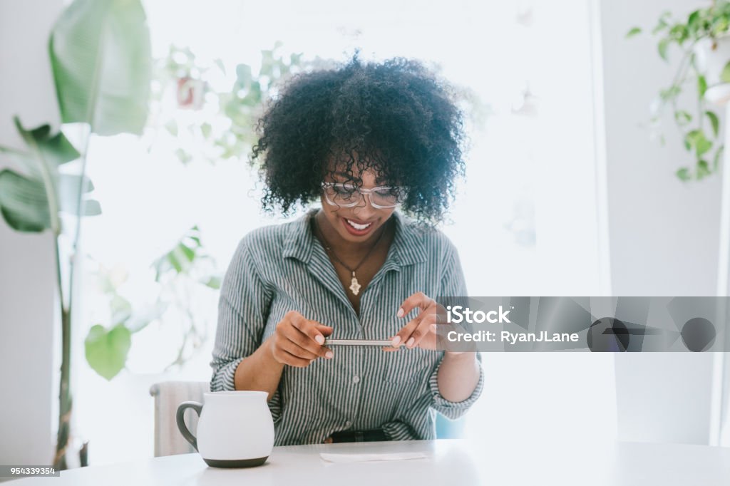 A Young Woman Does Remote Deposit Capture of Check A happy young adult woman enjoys time at her home, the house interior well designed and decorated with an assortment of interesting plants.  She does mobile banking from the convenience of her house, depositing a paycheck via Remote Deposit Capture. Down Payment Stock Photo