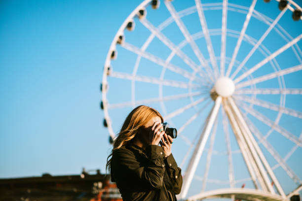 Female Film Photographer Explores Seattle Waterfront A woman walks on one of the piers of downtown Seattle, enjoying the setting sun and taking pictures with a film camera.  The Seattle ferris wheel is visible in the background. photographic film camera stock pictures, royalty-free photos & images
