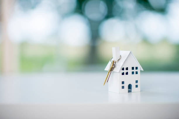 A pair of keys rests on a miniature model of a house, symbolizing a new home. A green tree and grass is seen in the background.