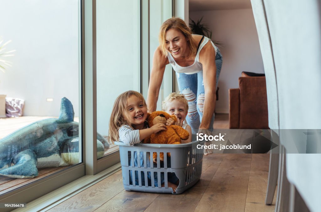 Mother playing with her children at home Happy young mother pushing children sitting in laundry basket. Mother and children playing at home. Mother Stock Photo