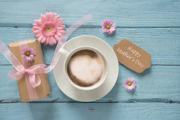 Coffee cup, pink flowers and a gift on pastel blue wood background with copy space, text Happy Mother's Day, top view from above