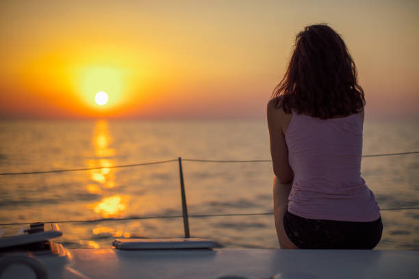 Mid adult woman sitting on sailboat deck at early morning and looking at sunrise and enjoying her first morning coffee. Yacht is on the move from one island to other