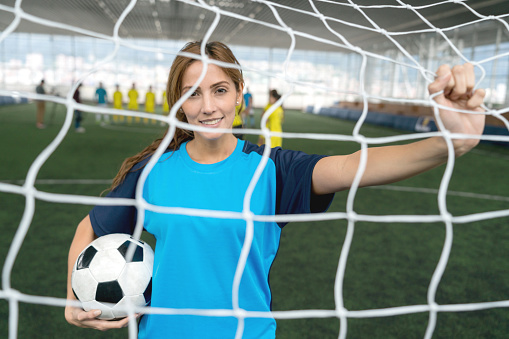 Beautiful female soccer player holding the net and a ball while looking at camera smiling with confidence