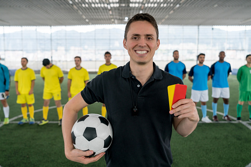 Portrait of friendly referee holding a yellow and red card looking at camera smiling very happy while te teams are standing behind