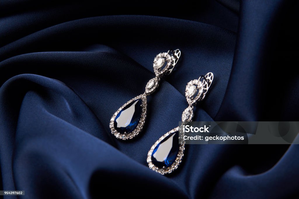Two Golden sapphire earrings with small diamonds Two Golden sapphire earrings with small diamonds. Pair of platinum earring with sapphire gemstone on blue satin background. Luxury female jewelry, close-up Jewelry Stock Photo
