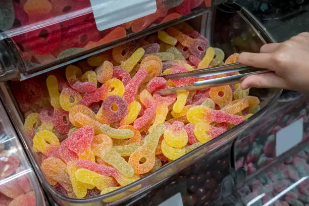 Photo of Kid hand picking fizzy and sour gummy jelly in plastic container with tong at confectionery shop.