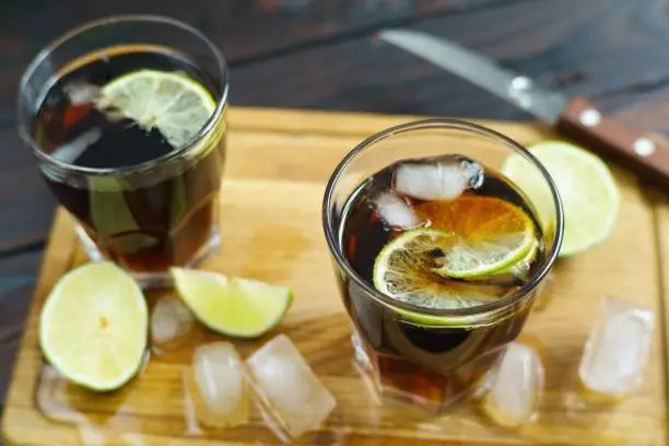 Refreshing iced longdrink Cuba Libre with rum and cola served on wooden board with lime slices and melting ice cubes