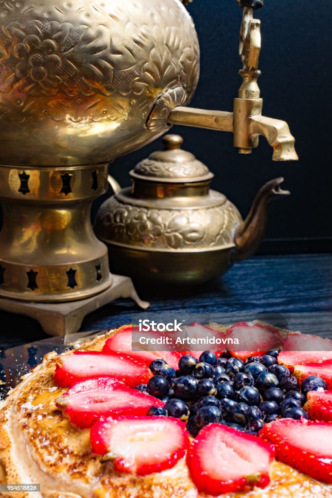 A stack of large pancakes cake in a metal plate and tea from a samovar on the background. close-up Large thin pancakes cake from strawberry, blueberry and condensed milk in a metal plate and tea from a old gold samovar and pot of tea on the black background. close-up Berry Stock Photo