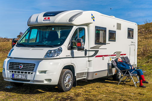 Bruddesta, Oland, Sweden - April 7, 2018: Travel documentary of everyday life and environment. Senior woman sitting outside Fiat Adria Coral S 670 SL camper enjoying the spring sun.