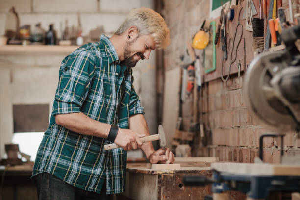 istock Attractive blonde young hipster bearded man by profession carpenter builder nailing wood board with hammer on a wooden table in the workshop. 954527730