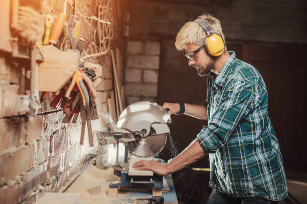 istock Young hipster bearded man with ear protectors by profession carpenter builder saws with a circular saw a wooden board on a wooden table in the workshop. 954528246