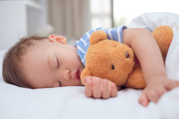 Cute healthy little Asian toddler baby boy child sleeping / taking a nap under blanket in bed while hugging teddy bear stock photo