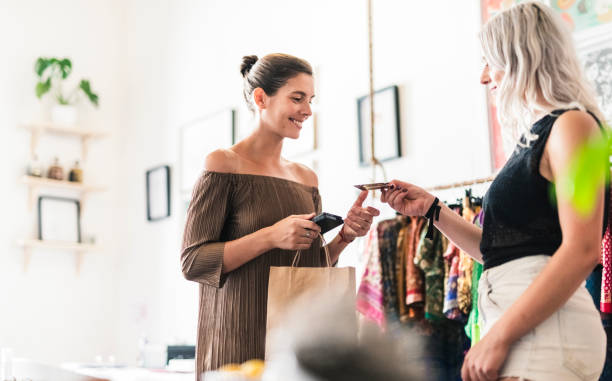 Woman doing credit card purchase from saleswoman Smiling woman doing credit card purchase. Saleswoman and customer are standing at clothing store. They are wearing casuals. clothing store credit card stock pictures, royalty-free photos & images