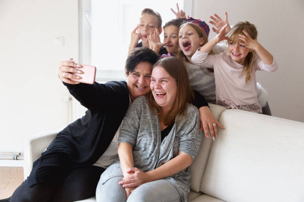 Kids photobombing their moms selfie Four children, aged seven to 12 years, photobomb the selfie their mothers  are trying to make. The mothers, two midadult women and best friends, are sitting on sofa, starting to make a selfie with cellphone, while their kids, 2 girls and 2 boys, are coming from behind and are making faces into the camera, everybody is having a lot of fun photo bomb stock pictures, royalty-free photos & images