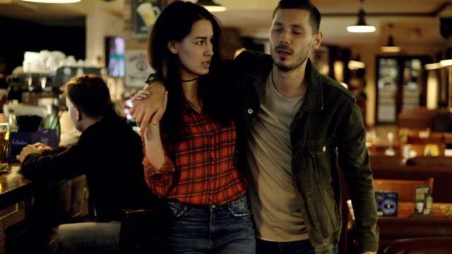 Young brunette is leading her drunk husband out of bar. Unshaven handsome young man is talking to his upset wife trying to explain himself. Alcoholism ruining family concept.