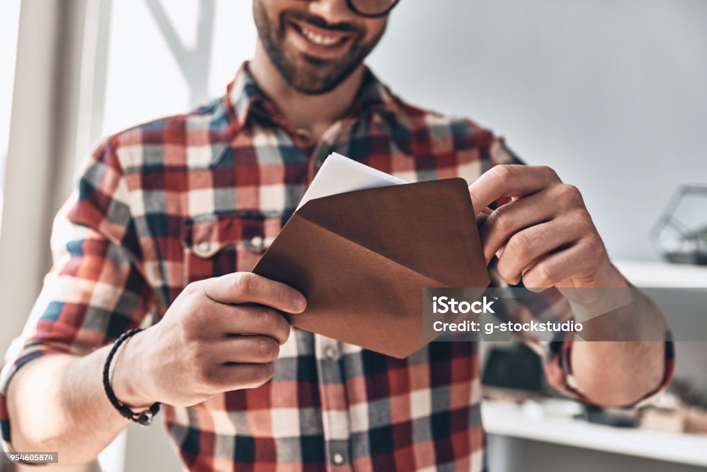 Receiving greeting card. Close up of young man opening envelope and smiling while standing indoors Opening Stock Photo
