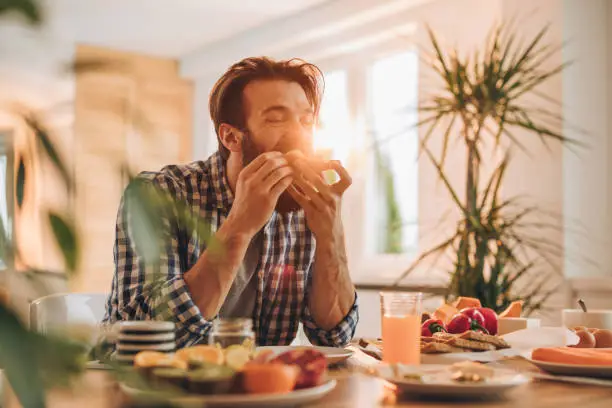 Young man eating sandwich for breakfast at dining table.