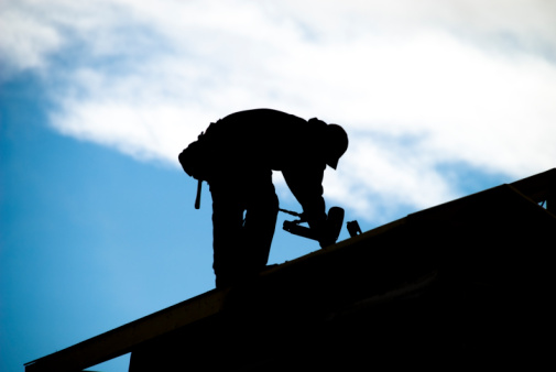 Silhouette of construction worker putting shingles on a house with a power nailer set against a blue sky.
