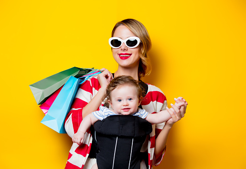 Mother and little baby in carrier have a shopping with bags. Models on yellow background