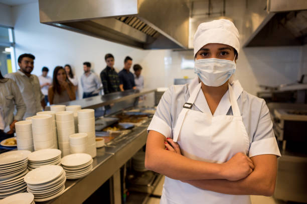 istock Female kitchen assistant working at a buffet restaurant wearing a protective mask and looking at camera 954735608