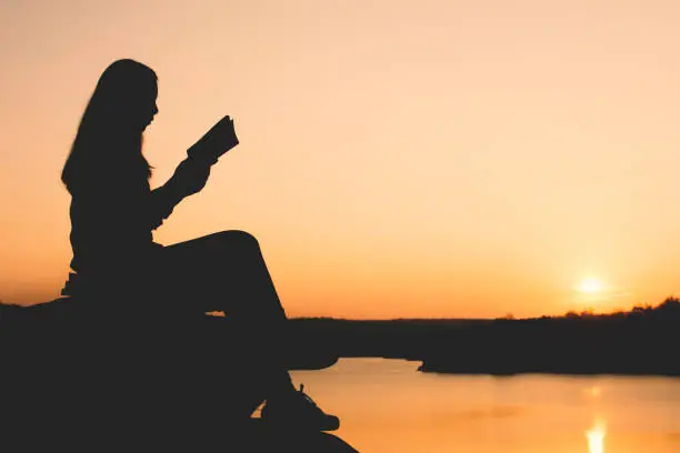 Photo of Silhouette Woman Reading Book While Sitting At Lakeshore During Sunset