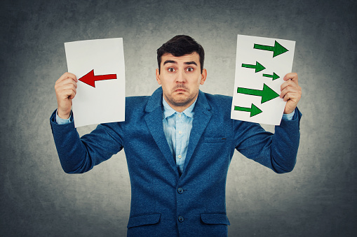 Confused young businessman holding two sheets with drawn arrows poinded to the left and right side. Undecided boy try to choose between two directions. Business decision and choice symbol., life perception concept.