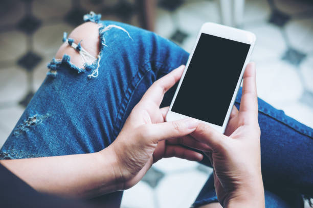 istock Mockup image of hand holding white mobile phone with blank black screen on thigh with a vintage tile floor in cafe , feeling relaxed 954896184