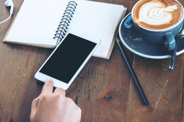 istock Mockup image of a hand touching on white mobile phone with blank black screen , hot coffee and vintage wooden table background 954896698