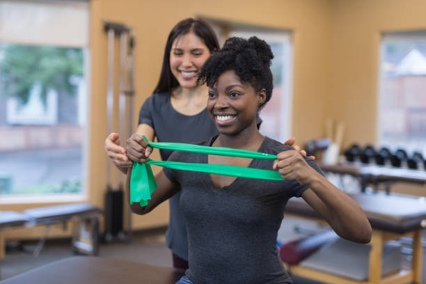 Physical therapy session with two women of color A physical therapist of Asian descent works with her patient, a millennial-age African American woman. They are doing a stretching exercise with a flexible exercise band. occupational therapy photos stock pictures, royalty-free photos & images