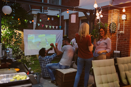 Friends watching sports on big screen in backyard. Drinking beer and cheering.