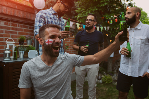 Friends watching sports on big screen in backyard. Man with English flag on face  cheering with beer.