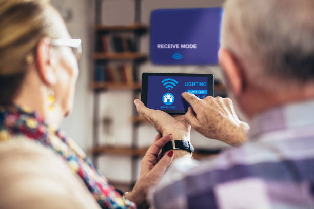 coppia di anziani seduta comodamente su un divano con la schiena che tiene il sistema di controllo remoto della casa su un tablet digitale - comfortably foto e immagini stock