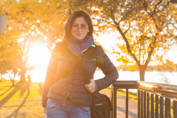 Woman with long brown hair is looking at camera while the sun is setting behind her on Charles river in a Boston urban park