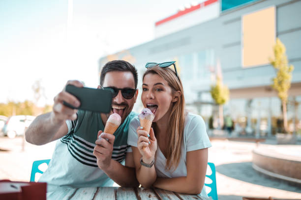 A beautiful young couple is taking selfie while they are sitting outdoors and having ice-creams in cornet stock photo