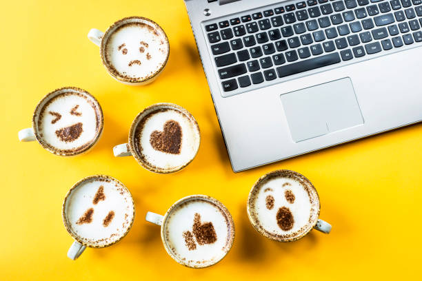 smile emoji painted on cups of cappuccino next to the laptop on a yellow background. emotions and communication online concept - tempera painting fotos imagens e fotografias de stock