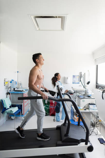 Handsome man doing a stress test looking very happy while walking on the treadmill Handsome man doing a stress test looking very happy while walking on the treadmill and black nurse at background looking at the cardiac monitor stress test stock pictures, royalty-free photos & images