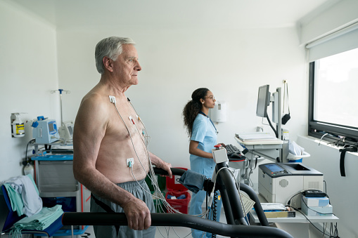 Senior man on a treadmill doing a stress test at the hospital while black nurse looks at the cardiac monitor very focused
